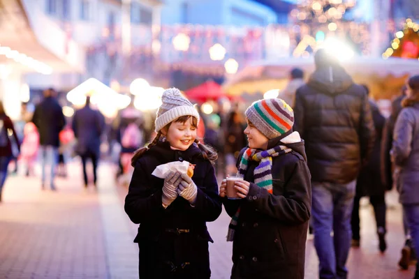 Linda niña y niño se divierten en el mercado tradicional de Navidad durante las fuertes nevadas. Niños felices comiendo salchichas tradicionales de curry llamadas wurst y bebiendo chocolate caliente. Gemelos amigos —  Fotos de Stock