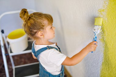 Sevimli küçük kız yeni evin rengiyle duvarı boyuyor. Aile onarım dairesi. Mutlu bebek duvarı boyuyor, paletle renk seçiyor, fırçayla eğleniyor, kapalı alanda.
