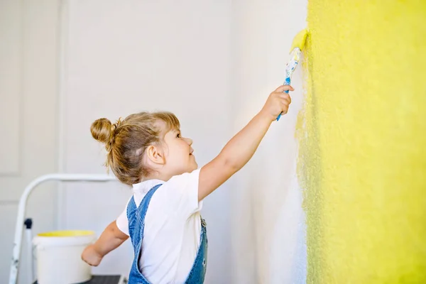 Pequeña niña divertida pintando la pared con color en una casa nueva. Casa de apartamento de reparación familiar. El niño feliz del bebé pinta la pared, eligiendo el color con la paleta, divirtiéndose con el cepillo, adentro —  Fotos de Stock