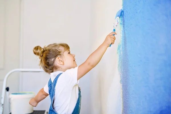 Zabawna dziewczynka malująca ścianę kolorem w nowym domu. Rodzinny dom remontowy. Szczęśliwe dziecko maluje ścianę, wybierając kolor z paletą, bawiąc się pędzlem, w pomieszczeniach — Zdjęcie stockowe