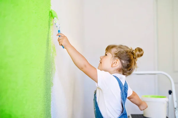 Pequeña niña divertida pintando la pared con color en una casa nueva. Casa de apartamento de reparación familiar. El niño feliz del bebé pinta la pared, eligiendo el color con la paleta, divirtiéndose con el cepillo, adentro —  Fotos de Stock