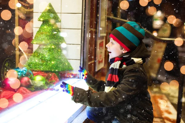 Cute little healthy school kid boy on Christmas market. Funny happy child in fashion winter clothes making window shopping decorated with gifts, xmas tree. Snow falling down, snowfall — Stock Photo, Image