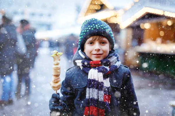 伝統的なドイツのクリスマスマーケットで串刺しにされた白いチョコレートを食べる小さなかわいい子供の男の子。雪の日にドイツの伝統的な家族市場で幸せな子供. — ストック写真