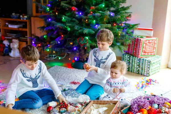 Dva malí kluci a roztomilá holčička zdobí vánoční stromeček se starými retro hračkami a míčky. Rodinná oslava rodinné dovolené před aparátem. Tři děti, bratři a sestra doma. — Stock fotografie