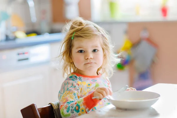 Schattig peutermeisje dat gezond pap eet van lepel als ontbijt. Schattig gelukkig baby kind in kleurrijke pyjama zitten in de keuken en leren met behulp van lepel. — Stockfoto