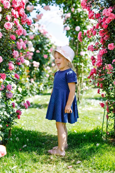 Porträt eines kleinen Mädchens im blühenden Rosengarten. Niedliche schöne schöne Kind Spaß mit Rosen und Blumen in einem Park an einem sonnigen Sommertag. Glücklich lächelndes Baby. — Stockfoto