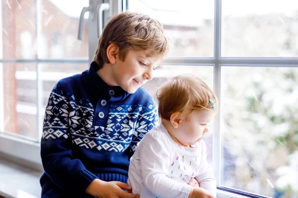 Glad bedårande pojke och söt liten flicka sitter nära fönstret och tittar ut på snö på juldagen eller morgonen. Leende barn, syskon, lillasyster och bror tittar på vintern snöfall. — Stockfoto