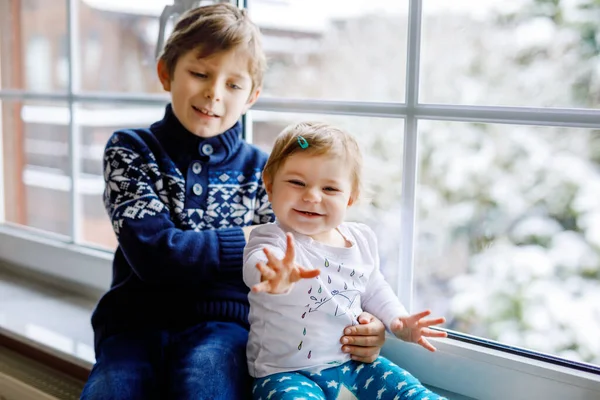 Niño adorable feliz y niña linda sentada cerca de la ventana y mirando afuera en la nieve en el día de Navidad o mañana. Niños sonrientes, hermanos, hermanita y hermano mirando las nevadas de invierno . — Foto de Stock