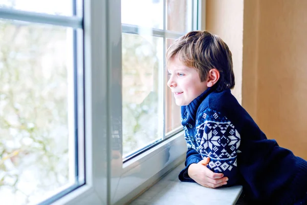 Glad bedårande liten pojke sitter nära fönstret och tittar ut på snö på juldagen eller morgonen. Leende friska barn fascinerad observera snöfall och stora snöflingor — Stockfoto
