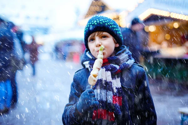 Niño lindo comiendo frutas cubiertas de chocolate blanco en pincho en el mercado tradicional alemán de Navidad. Niño feliz en el mercado familiar tradicional en Alemania durante el día nevado. — Foto de Stock