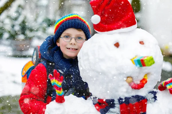 Renkli elbiseli, gözlüklü ve sırt çantalı küçük okul çocuğu ilkokul bittikten sonra kardan adamla eğleniyor. Çocuk kış günü karla oynuyor — Stok fotoğraf