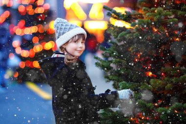 Little cute kid girl having fun on traditional Christmas market during strong snowfall. Happy child enjoying traditional family market in Germany. Schoolgirl standing by illuminated xmas tree. clipart