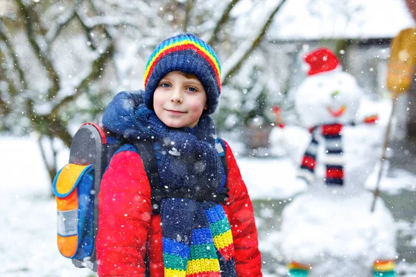 Little school kid boy in colorful clothes, with glasses and backpack having fun with snowman after elementary school end. Child playing with snow on winter day — Stock Photo, Image