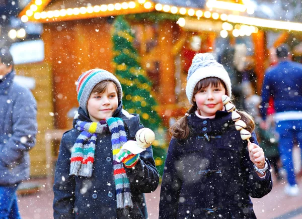 Little cute kid girl and boy eating white chocolate covered strawberries and apple on skewer on traditional German Christmas market. Happy children, best friends, twins and siblings on snowy day — Stock Photo, Image