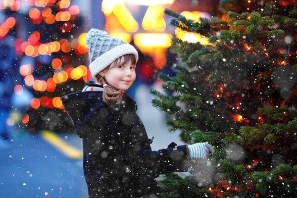 Niña linda que se divierte en el mercado tradicional de Navidad durante las fuertes nevadas. Niño feliz disfrutando del mercado familiar tradicional en Alemania. Colegiala de pie junto al árbol de Navidad iluminado. — Foto de Stock