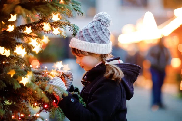 Little cute kid girl having fun on traditional Christmas market during strong snowfall. Happy child enjoying traditional family market in Germany. Schoolgirl standing by illuminated xmas tree. — Stock Photo, Image