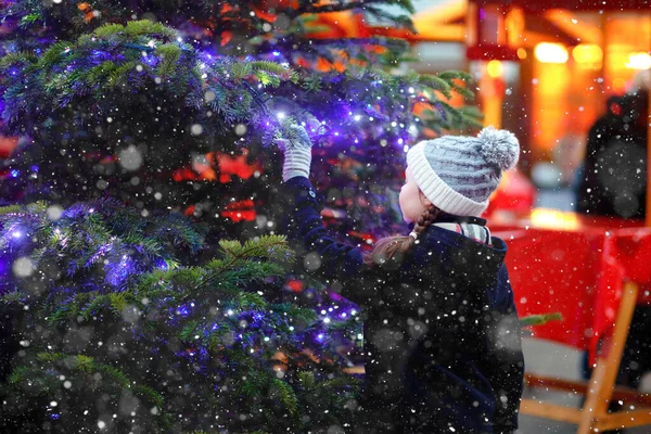 Niña linda que se divierte en el mercado tradicional de Navidad durante las fuertes nevadas. Niño feliz disfrutando del mercado familiar tradicional en Alemania. Colegiala de pie junto al árbol de Navidad iluminado. — Foto de Stock