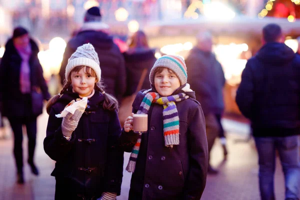 Cute little kids girl and boy having fun on traditional Christmas market during strong snowfall. Happy children eating traditional curry sausage called wurst and drinking hot chocolate. Twins friends — Stock Photo, Image