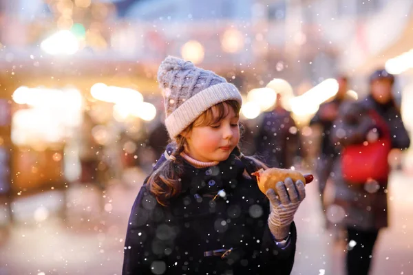 Cute little kid girl having fun on traditional Christmas market during strong snowfall. Happy child eating traditional curry sausage called wurst. schoolgirl standing by illuminated xmas tree. — Stock Photo, Image
