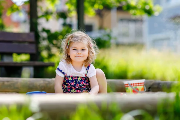 Menina bonito brincando na areia no parque infantil ao ar livre. Bebê bonito em calças vermelhas se divertindo no dia de verão quente ensolarado. Criança com brinquedos coloridos de areia. Bebê ativo saudável ao ar livre joga jogos — Fotografia de Stock