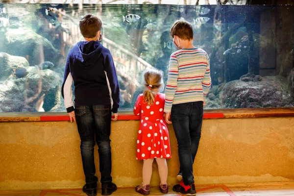 Two kids boys and toddler girl visiting together zoo aquarium. Three children watching fishes and jellyfishes. School boys wearing medicals masks due pandemic corona virus time. Family on staycation — Stock Photo, Image