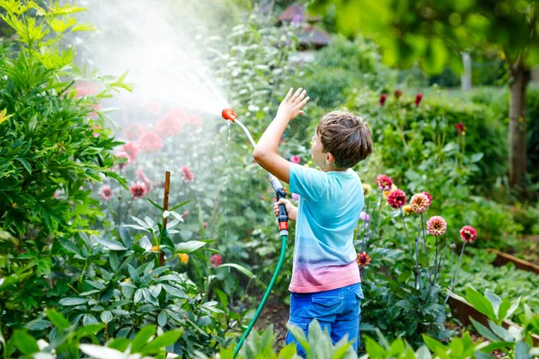 Belle petite école garçon arrosage des fleurs de jardin avec tuyau d'eau le jour d'été. Enfant heureux aidant dans le jardin familial, à l'extérieur, s'amusant avec éclaboussures — Photo