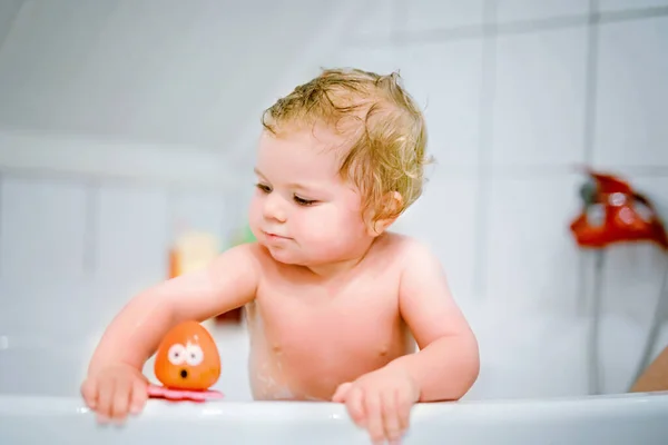 Cute adorable baby girl taking foamy bath in bathtub. Toddler playing with bath rubber toys. Beautiful child having fun with colorful gum toys and foam bubbles — Stock Photo, Image