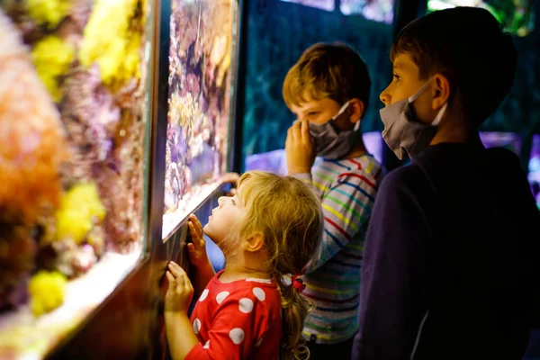 Två barn pojkar och småbarn flicka besöker tillsammans zoo akvarium. Tre barn tittar på fiskar och maneter. Skolpojkar bär medicinska masker på grund av pandemisk koronavirustid. Familj som stannar kvar — Stockfoto