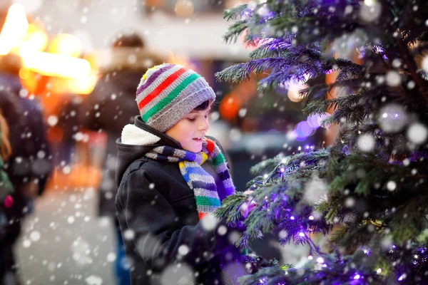 Little cute kid boy having fun on traditional Christmas market during strong snowfall. Happy child enjoying traditional family market in Germany. Schoolboy standing by illuminated xmas tree. — Stock Photo, Image