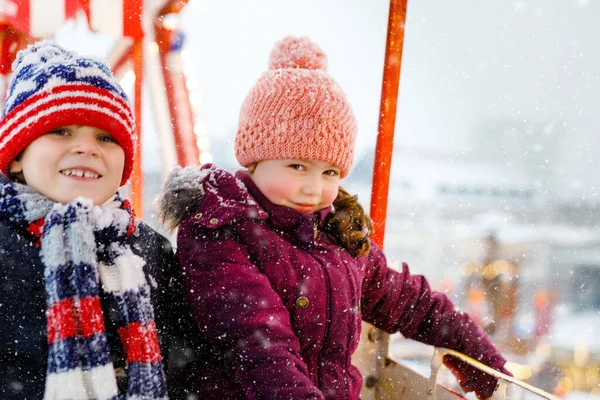 Duas crianças pequenas, menino e menina se divertindo na roda gigante no tradicional mercado de Natal alemão durante a forte queda de neve. Crianças felizes desfrutando do mercado familiar na Alemanha. Lindos irmãos.. — Fotografia de Stock