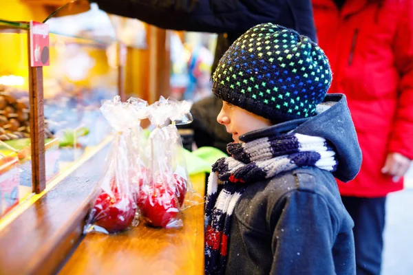 Kleine süße Junge in der Nähe von süßen Stand mit gezuckerten Äpfeln und Schokoladenfrüchten. Glückskind auf dem Weihnachtsmarkt in Deutschland. Traditionelle Freizeit für Familien an Weihnachten. Feiertag, Feier, Tradition. — Stockfoto