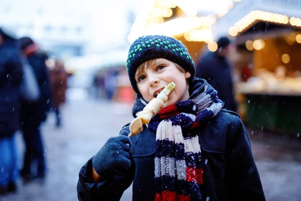 Pequeno menino bonito comendo frutas cobertas de chocolate branco no espeto no tradicional mercado de Natal alemão. Criança feliz no mercado familiar tradicional na Alemanha durante o dia nevado. — Fotografia de Stock