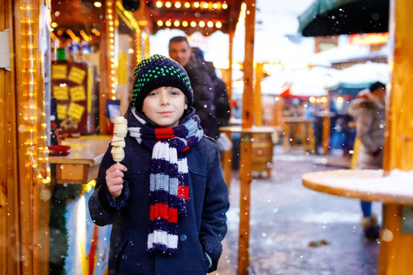 Der kleine süße Junge isst auf dem traditionellen deutschen Weihnachtsmarkt mit weißer Schokolade überzogene Früchte am Spieß. Glückliches Kind bei verschneitem Tag auf traditionellem Familienmarkt in Deutschland. — Stockfoto