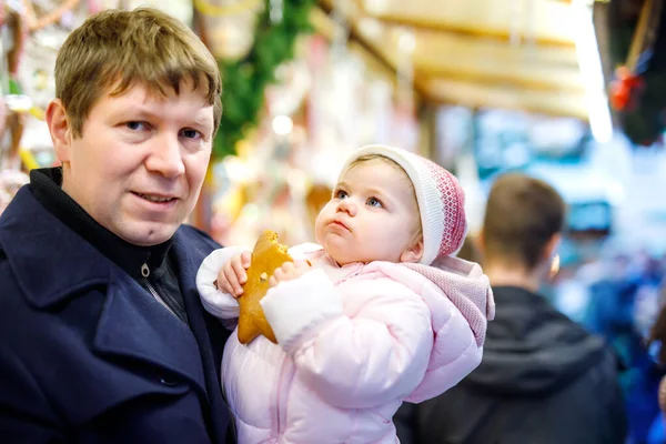 Middle aged father holding baby daughter near sweet stand with gingerbread and nuts. Happy family on Christmas market in Germany. Cute girl eating cookie called Lebkuchen. Celebration xmas holiday. — Stock Photo, Image