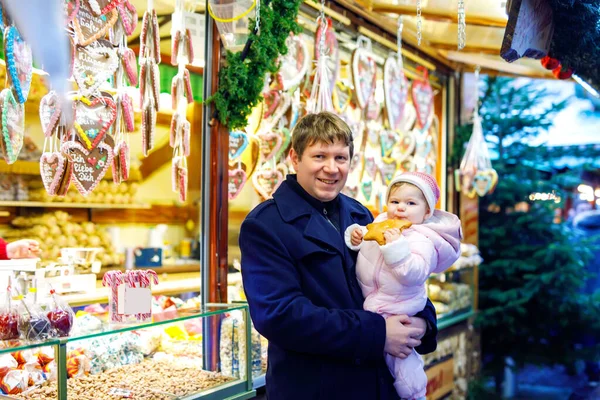 Middle aged father holding baby daughter near sweet stand with gingerbread and nuts. Happy family on Christmas market in Germany. Cute girl eating cookie called Lebkuchen. Celebration xmas holiday. — Stock Photo, Image