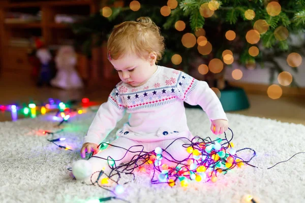 Schattige baby meisje met kleurrijke lichten bloemenslinger in schattige handen. Klein kind in feestelijke kleding versieren kerstboom met familie. Eerste viering van de traditionele feestdag genaamd Weihnachten — Stockfoto