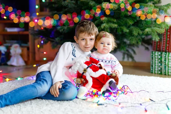 Entzückende kleine Mädchen und Bruder mit bunten Lichtergirlanden und Spielzeug-Weihnachtsmann in niedlichen Händen. — Stockfoto