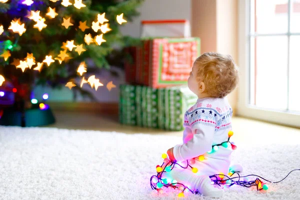 Schattige baby meisje met kleurrijke lichten bloemenslinger in schattige handen. Klein kind in feestelijke kleding versieren kerstboom met familie. Eerste viering van de traditionele feestdag genaamd Weihnachten — Stockfoto