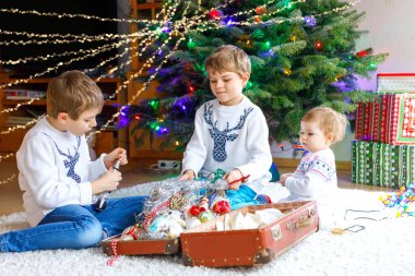 İki küçük oğlan ve sevimli kız bebek Noel ağacını eski model oyuncaklar ve toplarla süslüyorlar. Aile tatili için aile hazırlık kutlaması. Evde üç çocuk, erkek ve kız kardeş var..