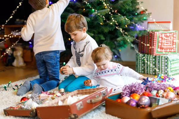 Zwei kleine Jungen und ein entzückendes Mädchen schmücken den Weihnachtsbaum mit altem Spielzeug und Bällen. Familienvorbereitungsfeier des Familienurlaubs. Drei Kinder, Geschwister zu Hause. — Stockfoto