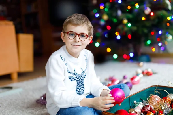 Beautiful kid boy with eye glasses and colorful vintage xmas toys and balls in old suitcase. Child decorating Christmas tree — Stock Photo, Image