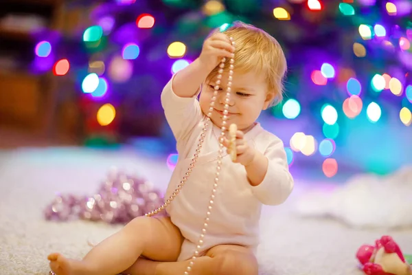 Schattige baby meisje met kleurrijke lichten bloemenslinger in schattige handen. Klein kind in feestelijke kleding versieren kerstboom met familie. Eerste viering van de traditionele feestdag genaamd Weihnachten — Stockfoto