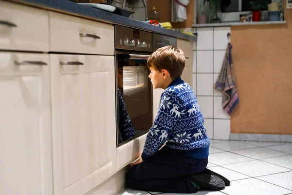 Cute funny blond kid boy baking muffins in domestic kitchen. Child having fun with helping, sitting near ofen and waiting for cupcakes — Stock Photo, Image