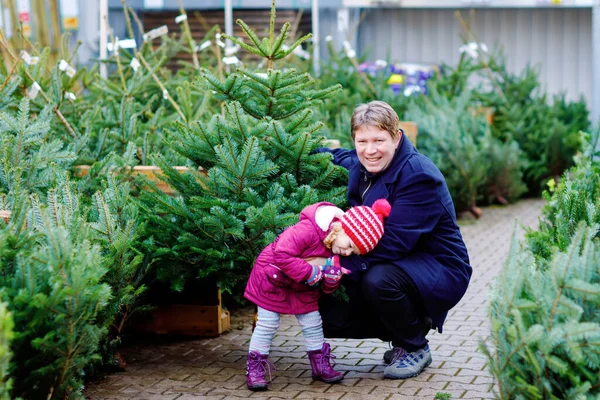 Kleines Mädchen und Vater mit Weihnachtsbaum auf einem Markt. Glückliche Familie, niedliche Baby-Tochter und Mann mittleren Alters in Wintermode Kleidung auswählen und kaufen Weihnachtsbaum im Outdoor-Shop. — Stockfoto