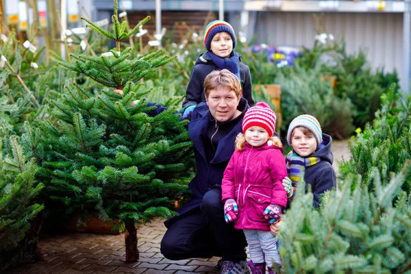 Маленькая девочка, два мальчика и отец держат рождественскую елку на рынке. Счастливая семья, милые дети и мужчина средних лет в зимней модной одежде, выбирающий и покупающий новогоднюю елку в магазине под открытым небом. — стоковое фото