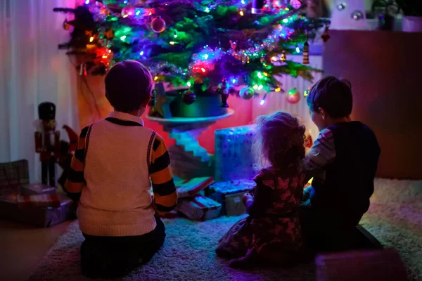 Dos niños pequeños y un niño adorable sentado junto al árbol de Navidad con viejos juguetes vintage y bolas con un montón de regalo en la víspera de Navidad. Preaparación familiar celebración de la fiesta familiar —  Fotos de Stock