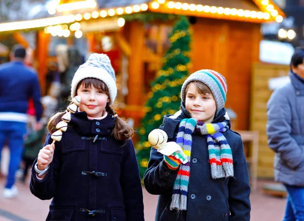 Malá roztomilá holčička a chlapec jíst bílou čokoládu pokryté jahody a jablko na špejli na tradičním německém vánočním trhu. Happy children, best friends, twins and siblings on snowy day — Stock fotografie