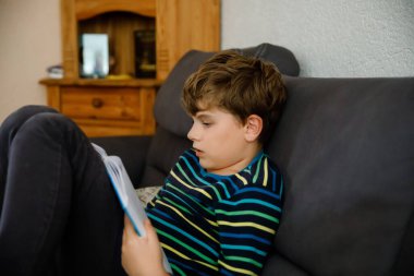 Şirin sarışın çocuk dergi ya da kitap okuyor. Heyecanlı çocuk sesli okuyor, kanepede oturuyor. Okul çocuğu, aile, eğitim. Covid zamanı evde eğitim