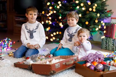 İki küçük oğlan ve sevimli kız bebek Noel ağacını eski model oyuncaklar ve toplarla süslüyorlar. Aile tatili için aile hazırlık kutlaması. Evde üç çocuk, erkek ve kız kardeş var..