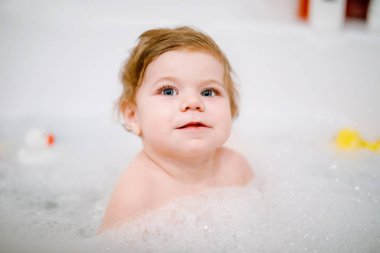 Küvette köpüklü banyo yapan sevimli, sevimli bir kız. Bebek banyo lastiği oyuncaklarıyla oynuyor. Güzel çocuk renkli sakız oyuncakları ve köpüklerle eğleniyor.